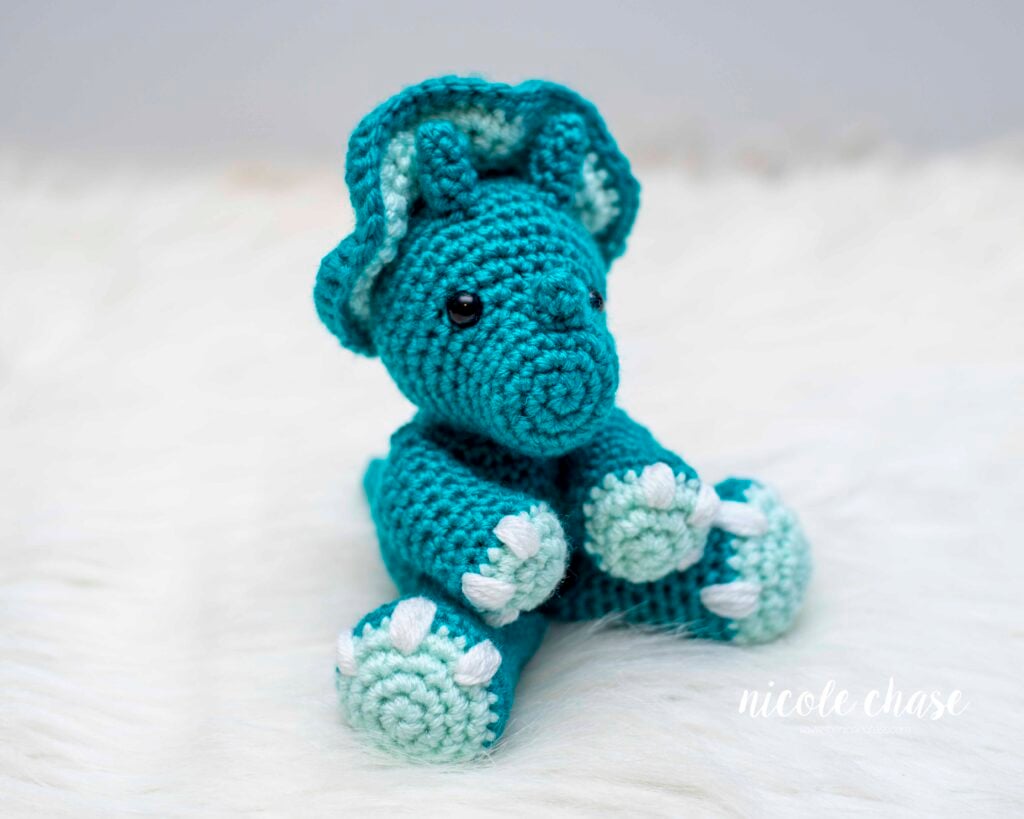 A small, teal crochet dinosaur sitting on a white faux fur. The dinosaur features a large frill, three horns, and white nails. 