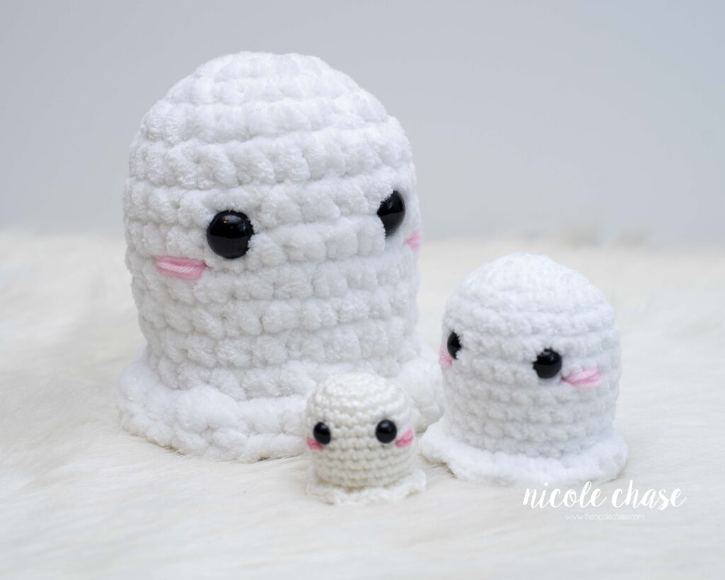 Three white crocheted beginner ghosts in varying sizes and yarn textures. The larges and medium ghosts are made with thick, fuzzy plush, while the smallest is made with a smooth acrylic yarn. All feature black safety eyes and pink embroidered cheeks. 
