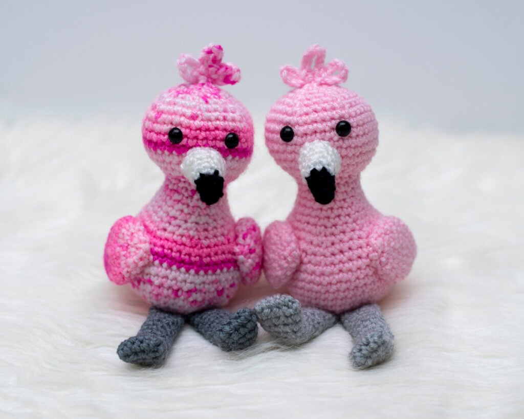 Two crocheted amigurumi flamingos, one made with solid light pink yarn and the other with a variegated pink and white yarn, sitting on a white faux fur surface. This image showcases the free flamingo crochet pattern worked in different yarn styles to highlight texture and color variations.