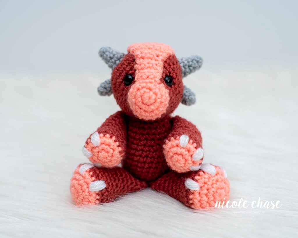 A close-up of Mini Archie the Ankylosaurus amigurumi, a small crochet dinosaur in dark red and peach yarn with gray horns, safety eyes, and white nails, sitting posed on a soft white background.