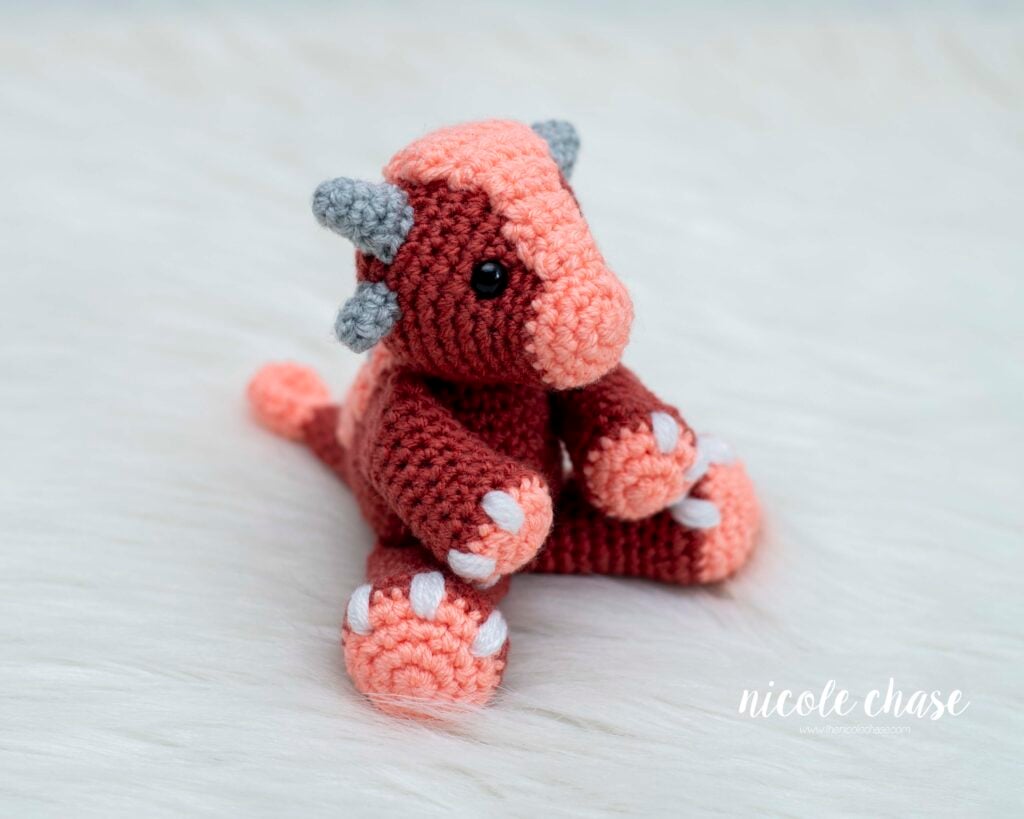 Mini Archie the Ankylosaurus amigurumi, a small crochet dinosaur in shades of red/pink with gray horns and white toes, sitting on a white fluffy surface.