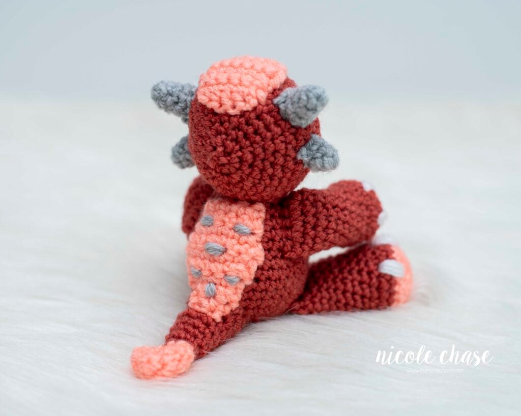 Back view of Mini Archie the Ankylosaurus amigurumi, a small crochet dinosaur, showing the club tail, textured back armor/plates in gray and peach yarn, and small gray horns on its head.