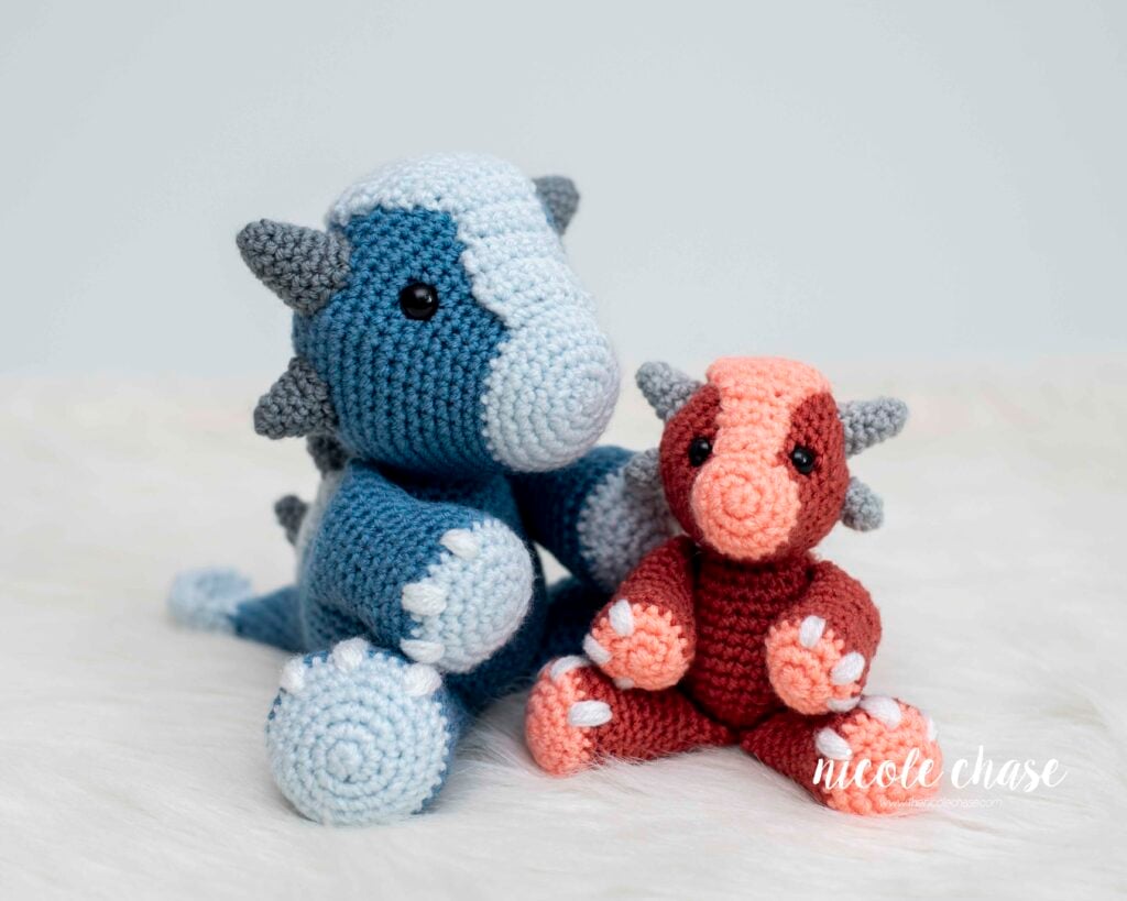 Mini Archie the Ankylosaurus (right, in red and peach) sitting next to the original, larger Archie the Ankylosaurus (left, in two shades of blue), showcasing the size difference between the small dinosaur crochet pattern and the full-size amigurumi pattern.