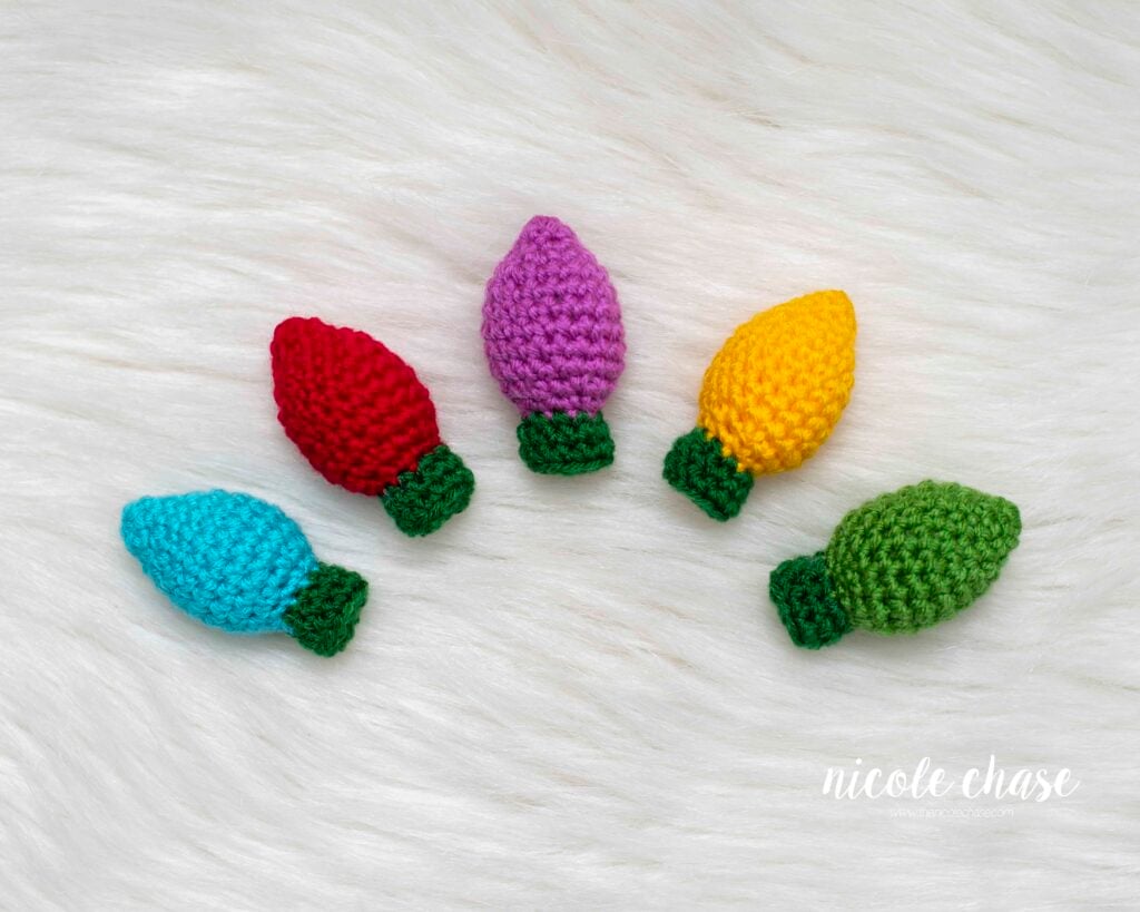 Five finished, colorful crochet Christmas light bulbs (blue, red, purple, yellow, and green) lying individually on a white fluffy surface. These are the finished components for the free crochet Christmas lights pattern. 