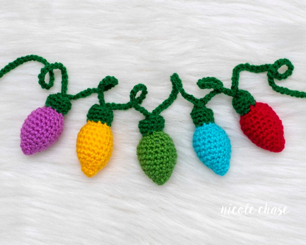 Five colorful crochet Christmas lights (purple, yellow, green, blue, red) connected by a green chain, arranged as a garland on a white fluffy surface. Example of the finished FREE crochet Christmas lights pattern. 