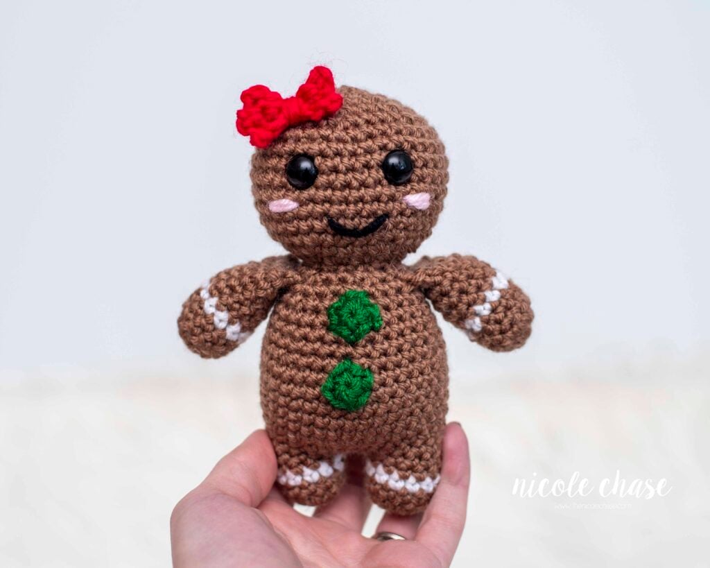 Finished Gerry the Gingerbread Girl amigurumi held in a hand, customized with a red hair bow and green buttons. This is the FREE crochet gingerbread man pattern, a quick, nearly no-sew project. 