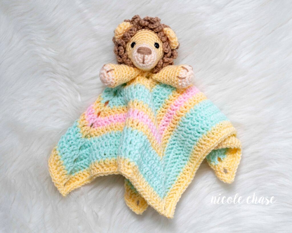Finished Hug Me Lion Lovey crochet security blanket lying on a white background, featuring a pastel striped blanket in yellow, pink, and mint green.
