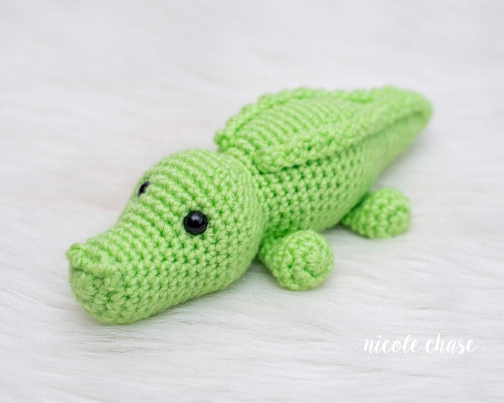A finished amigurumi alligator made with bright green yarn, featuring black safety eyes and textured ridges along its back. The free alligator crochet pattern is styled on a white textured surface.