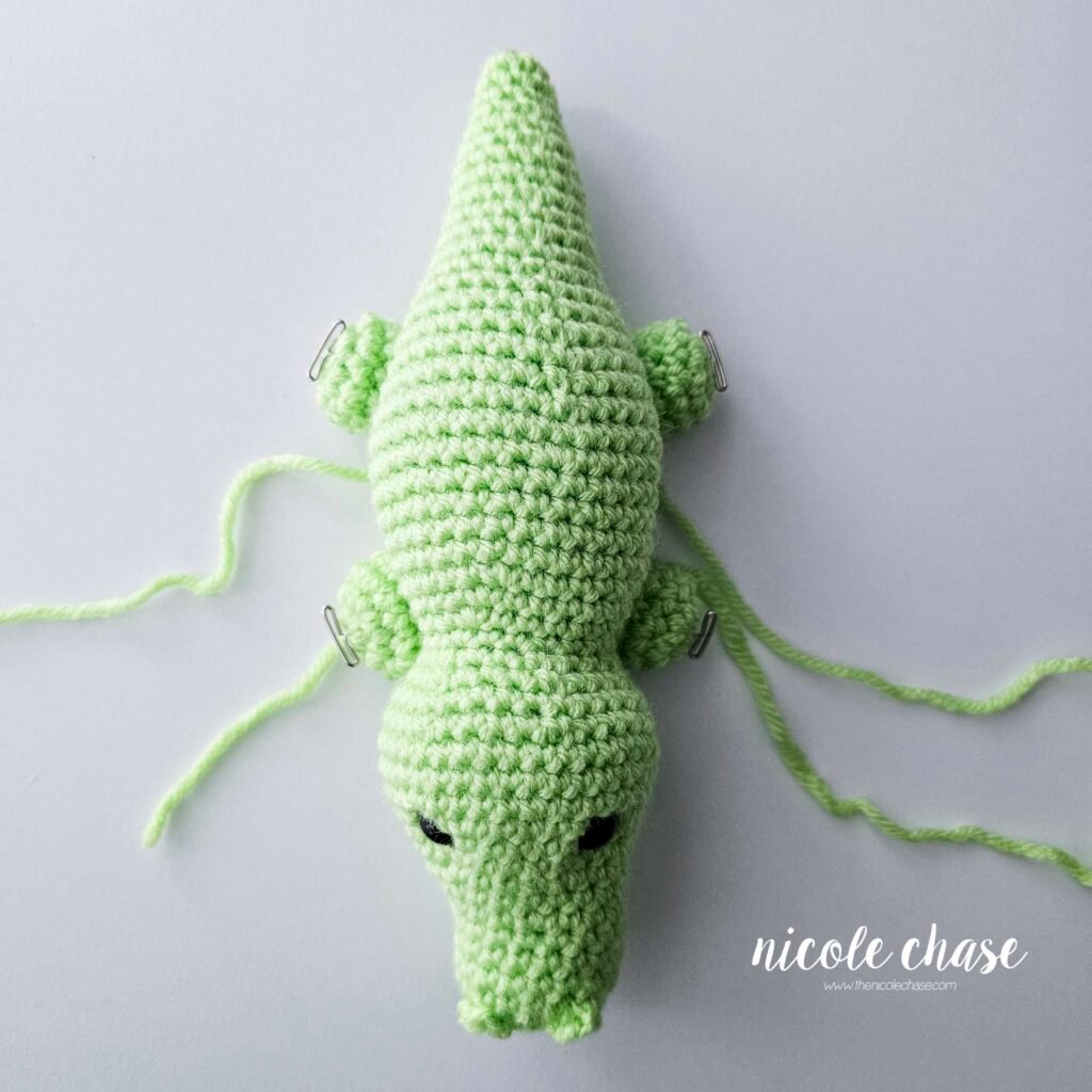 The legs of the crochet alligator have been pinned into place on the bottom sides of the body, view 1.