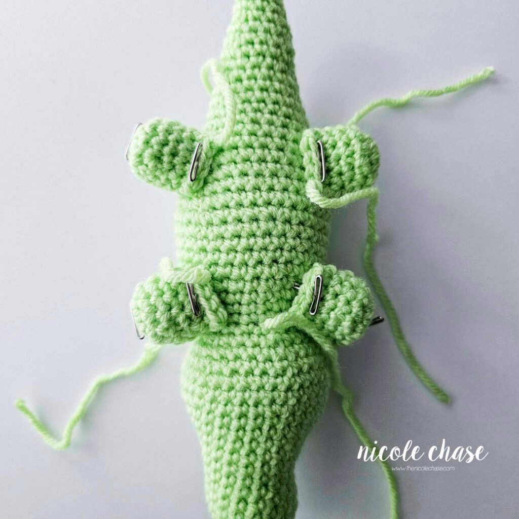 The legs of the crochet alligator have been pinned into place on the bottom sides of the body, view 2.