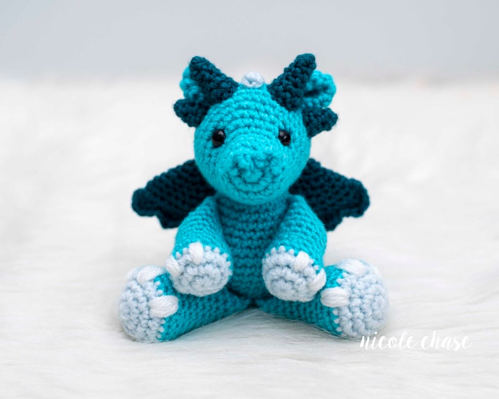 Face on front view of the free dragon crochet pattern for Danny the Dragon. The dragon is made of bright blue yarn with dark teal wings and spikes and light blue hands and feet.