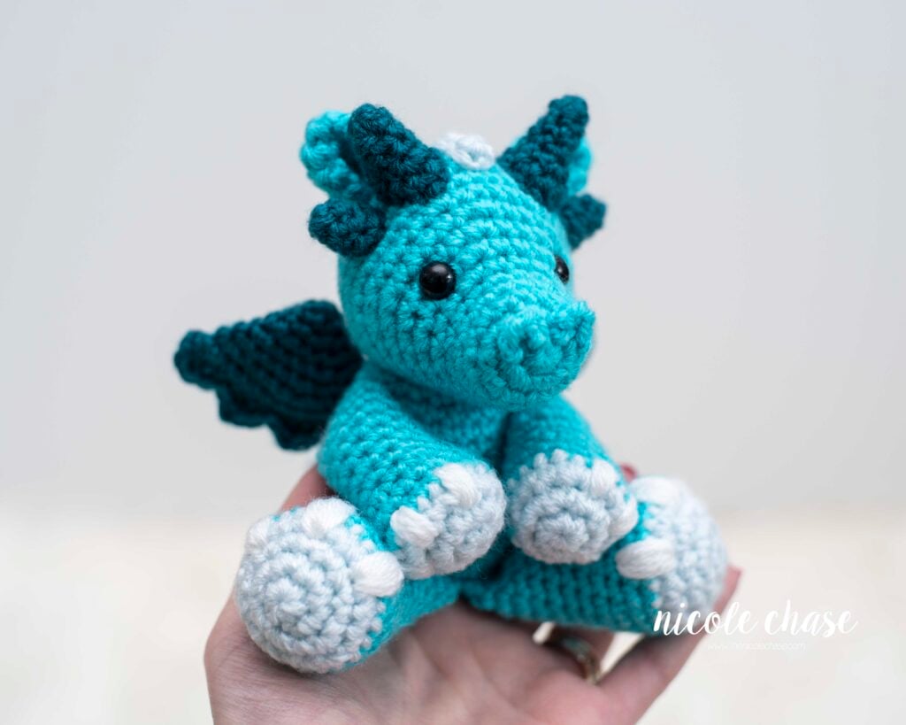 A hand holding a finished turquoise and teal amigurumi dragon to show its small portable size. Danny the Dragon features teal wings and horns, with light blue feet. This photo demonstrates the scale of the finished project from the free dragon crochet pattern.
