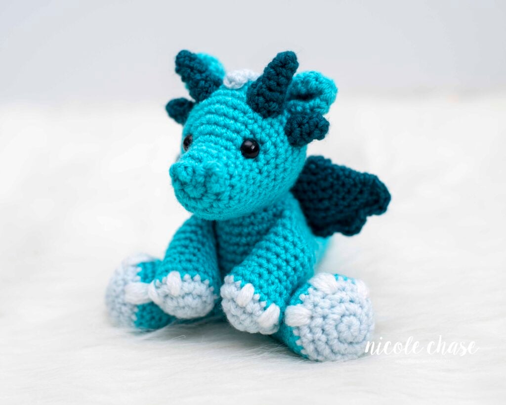 A vibrant turquoise and teal crochet dragon (Danny the Dragon) sitting on a white fluffy surface. The dragon has black safety eyes, 4 teal horns, small turquoise ears, and two teal wings. It features light blue feet. Made with this free dragon crochet pattern.