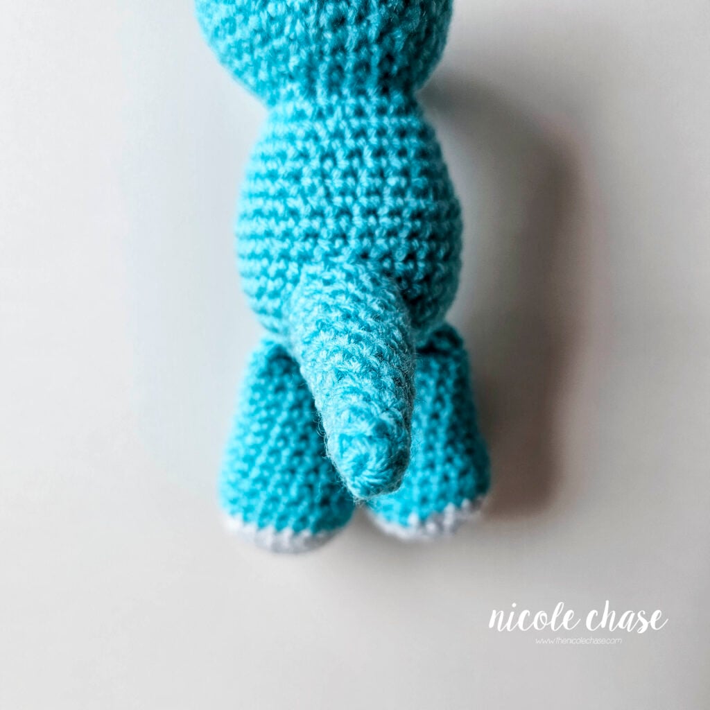 The crochet dragon's tail has been sewn into place on the back of the body, view from the back with the dragon lying face down.