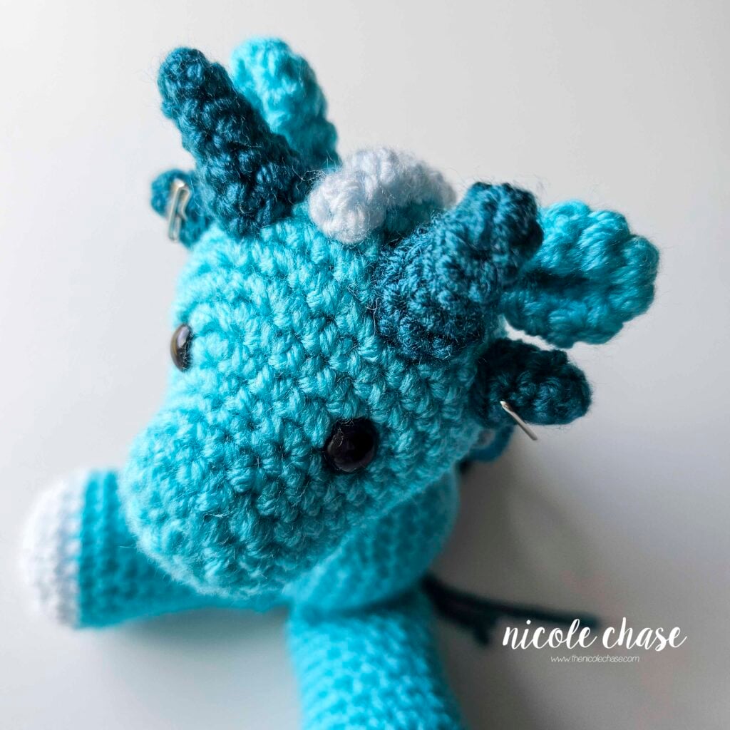 The small teal spikes of the crochet dragon have been pinned into place on each side of the dragon's head, just under the large spikes, and are awaiting sewing.