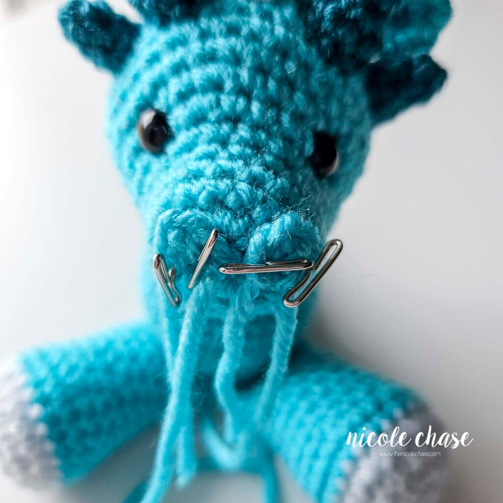 The nostrils have been pinned into an upside down U shape on the front of the crochet dragon's head and are awaiting sewing.