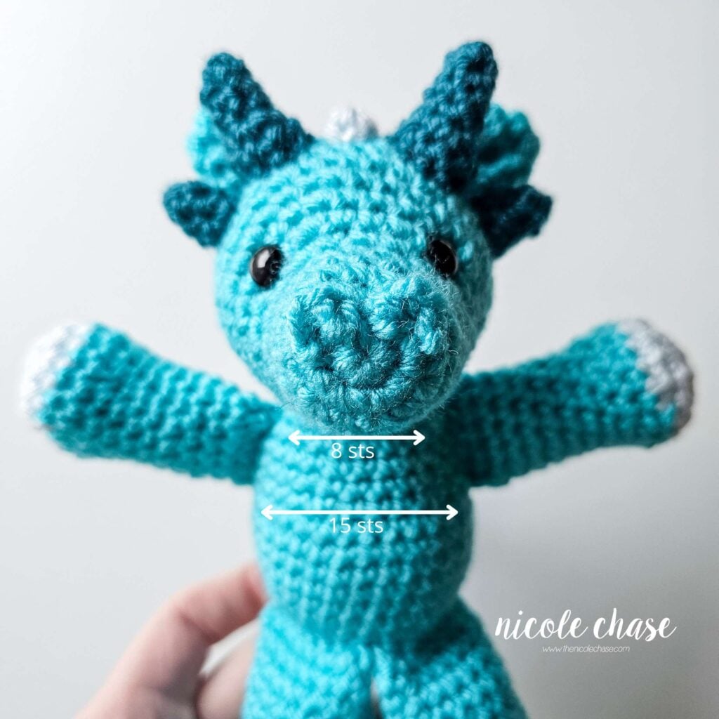 The dragon's crochet arms have been sewn into place on each side of the body. The dragon's arms are spread wide, and a graphic is detailing the stitch spacing between each arm.