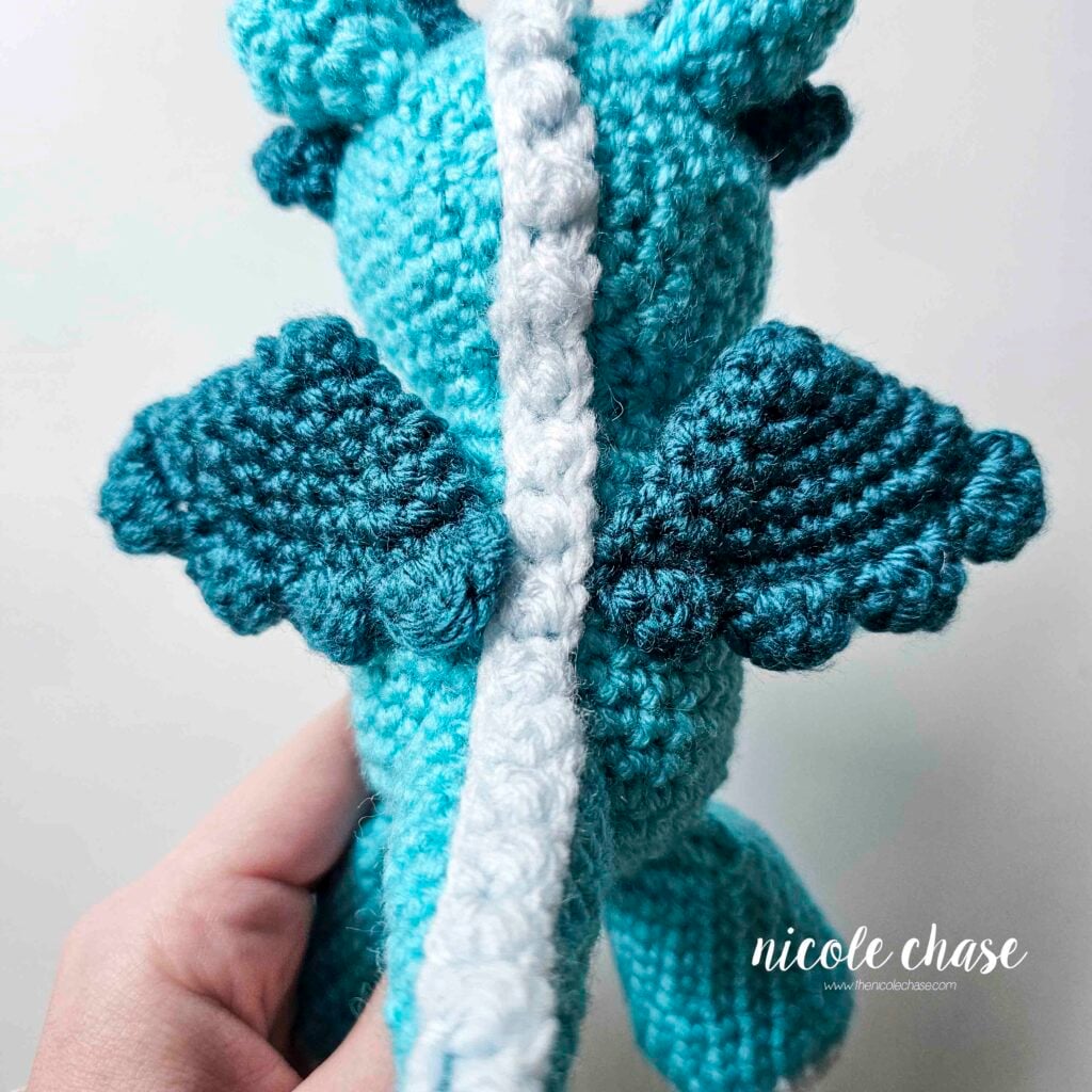 A view of the back of the crochet dragon showing the teal wings on each side of the body, with the ridge going down in between.