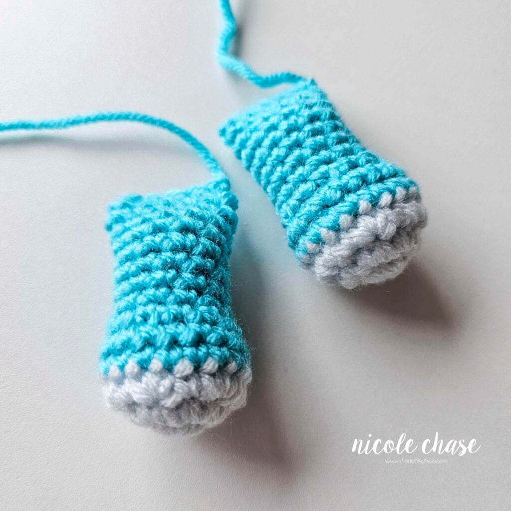 Two finished crochet arms for the dragon amigurumi made in bright blue yarn with light blue tips. The arms are filled with stuffing, crocheted closed, and awaiting assembly.