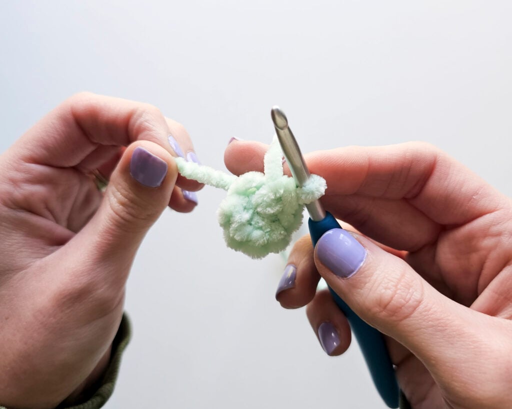 Two hands on a white background are holding a crochet hook with a circle of light green stitches, after just finishing the magic ring alternative method using plush chenille yarn to prevent breakage.