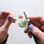 Two hands on a white background are holding a crochet hook with a circle of light green stitches, after just finishing the magic ring alternative method using plush chenille yarn to prevent breakage.