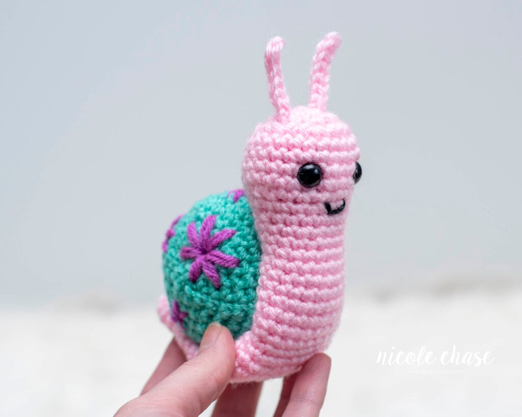 Side view of a crocheted amigurumi snail held in a hand, showcasing the teal shell with purple embroidered floral details. The snail is made with pink yarn and features a simple friendly face with black eyes.