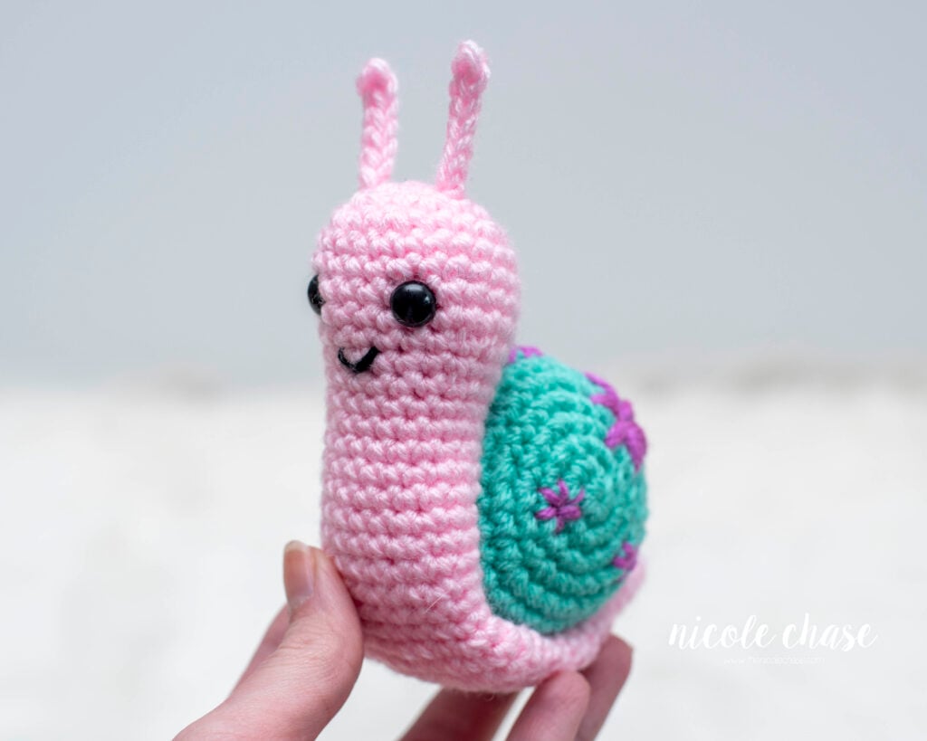A small pink crocheted amigurumi snail named Shell held in a hand. The snail has a teal shell decorated with small embroidered purple flowers, black safety eyes, a stitched smile, and two small antennae. Made with this free snail crochet pattern.