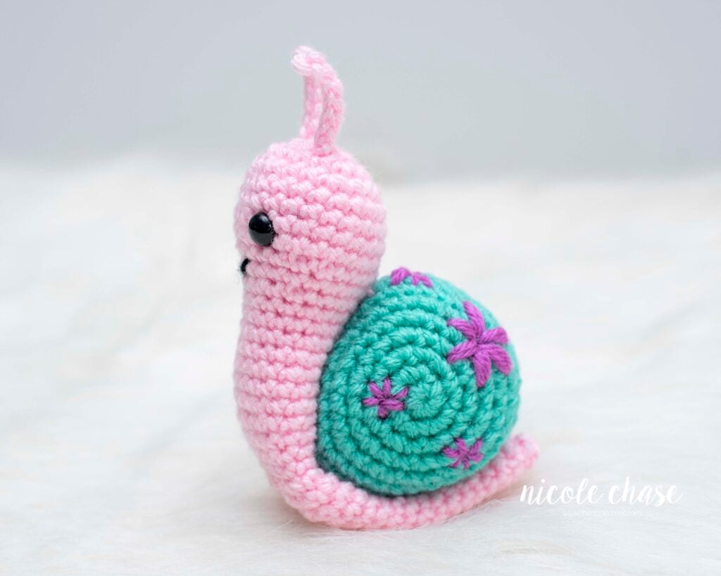 A side profile of Shelly the snail, a pink crocheted amigurumi sitting on a white textured surface. The teal snail shell is decorated with several purple embroidered flowers of varying sizes .The snail has black safety eyes and small pink antennas.