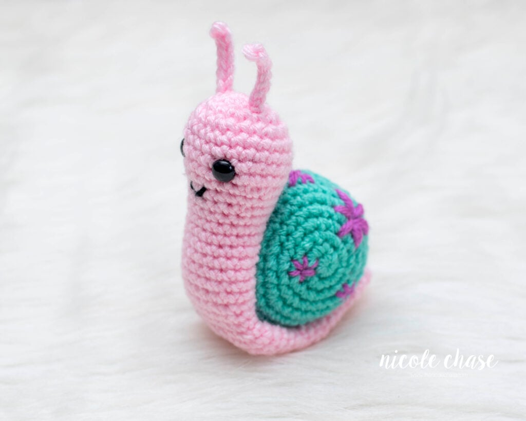 A small pink crocheted amigurumi snail, Shelly, sitting on a white fuzzy surface. The snail features a teal shell with purple embroidered flowers and is shown from an angle, highlighting its rounded head, black safety eyes and small antennae.