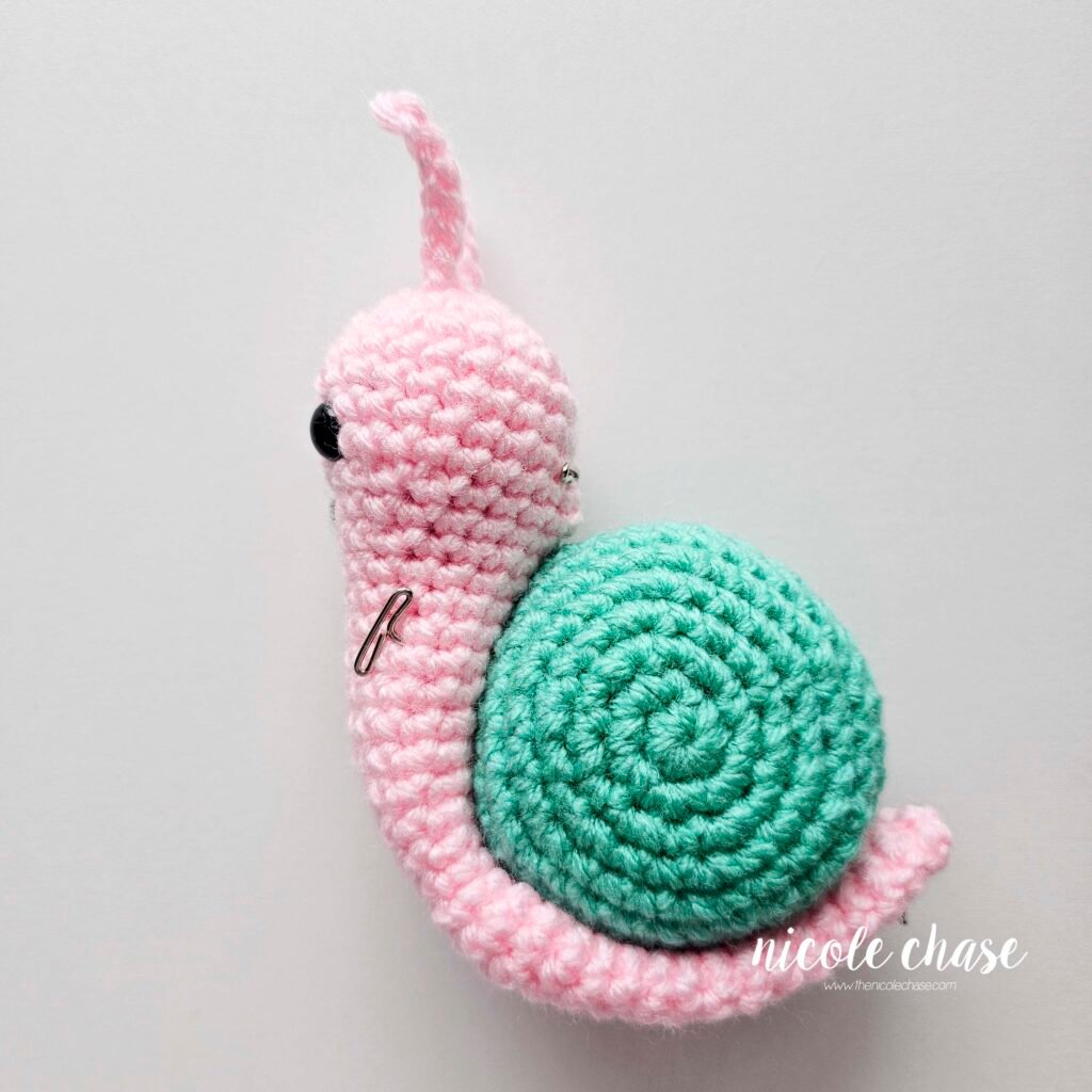Pinning the crochet snail's pink body in place curled around the teal shell prior to sewing, side view.