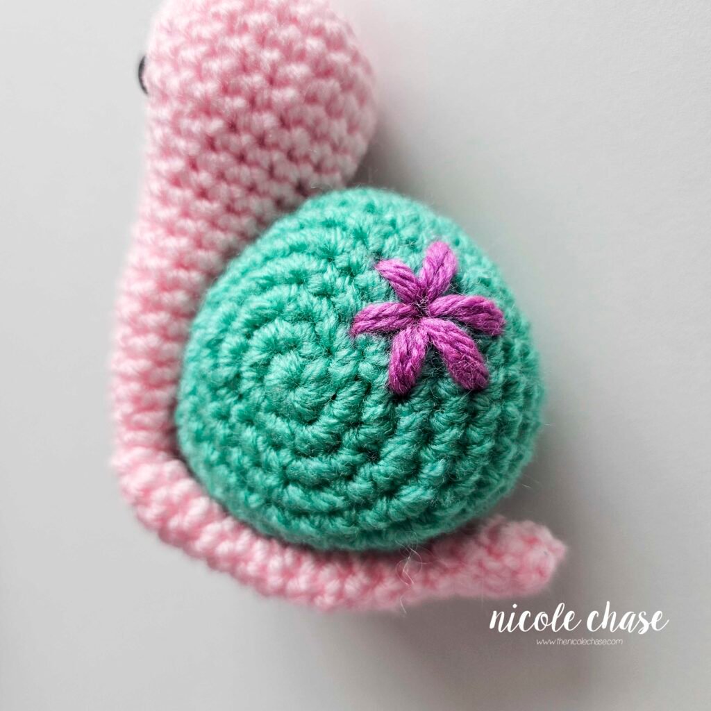 The finished embroidered large flower in purple yarn on the teal shell of the free snail crochet pattern.