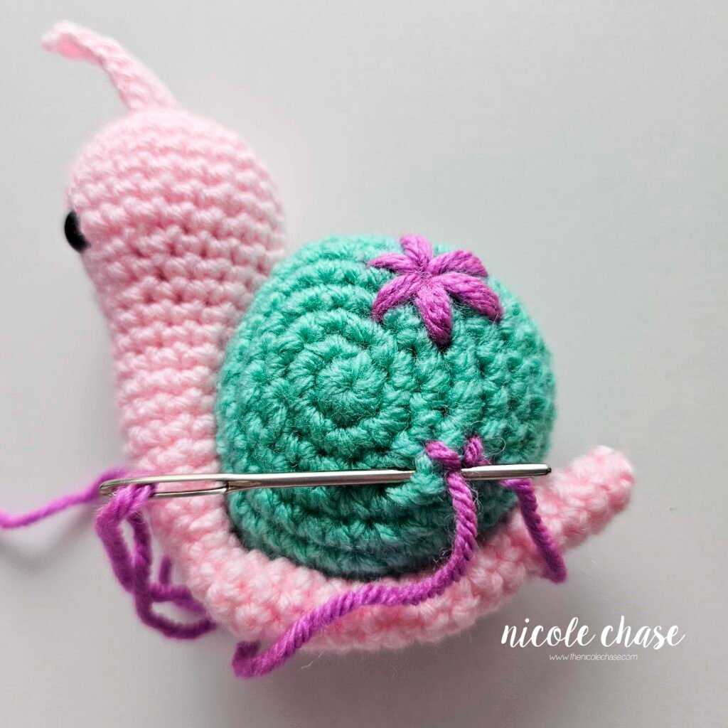 Embroidering a small flower on the shell of the crochet snail with purple yarn, step 4.