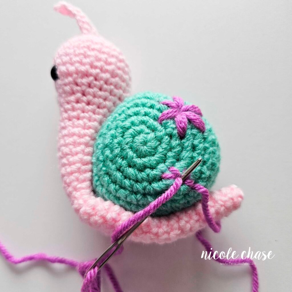Embroidering a small flower on the shell of the crochet snail with purple yarn, step 5.