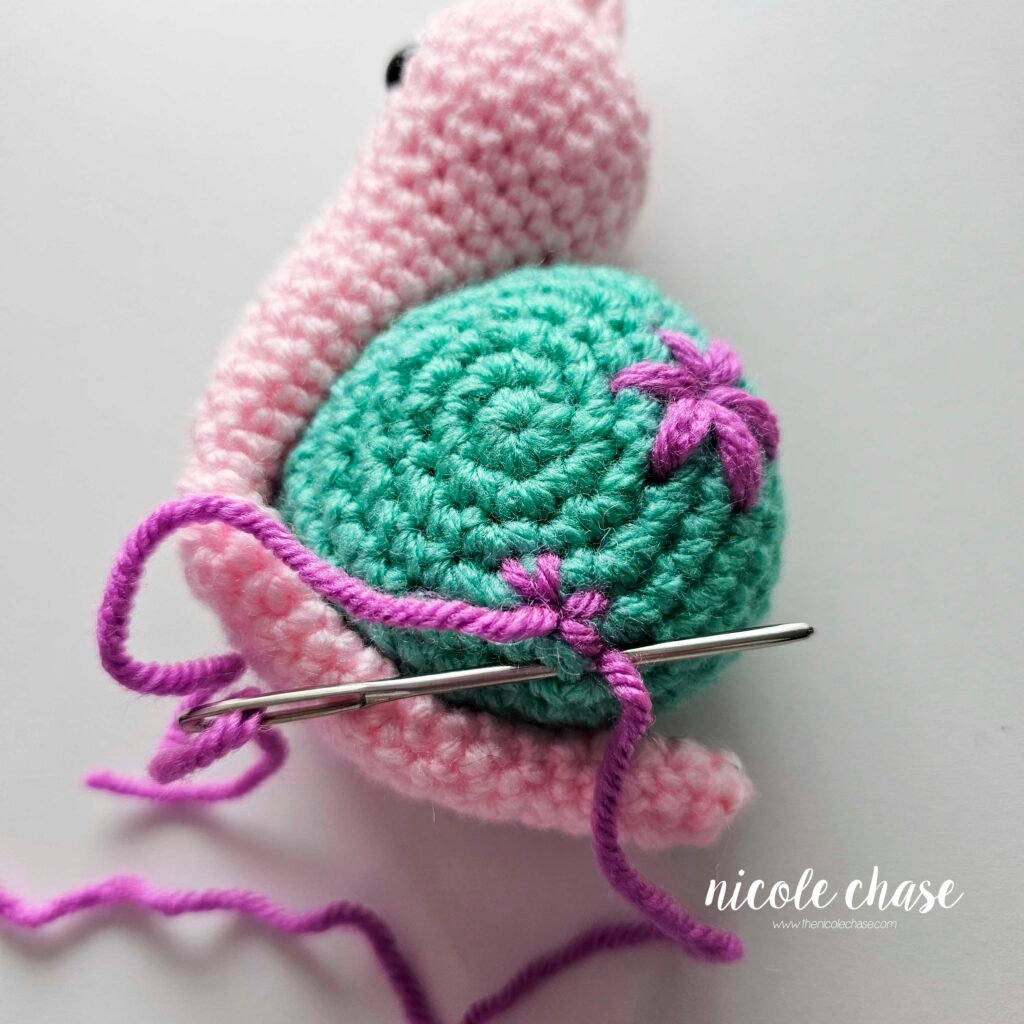 Embroidering a small flower on the shell of the crochet snail with purple yarn, step 6.