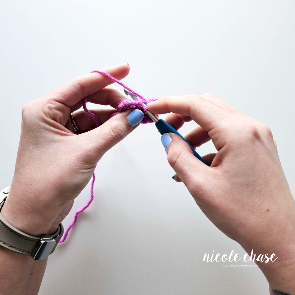 Close-up view of hands holding a blue ergonomic crochet hook using the knife grip style. The hook is inserted into the stitch, and the tension hand, guided by the left index finger, holds the purple yarn taught, demonstrating how to control the working loop.
