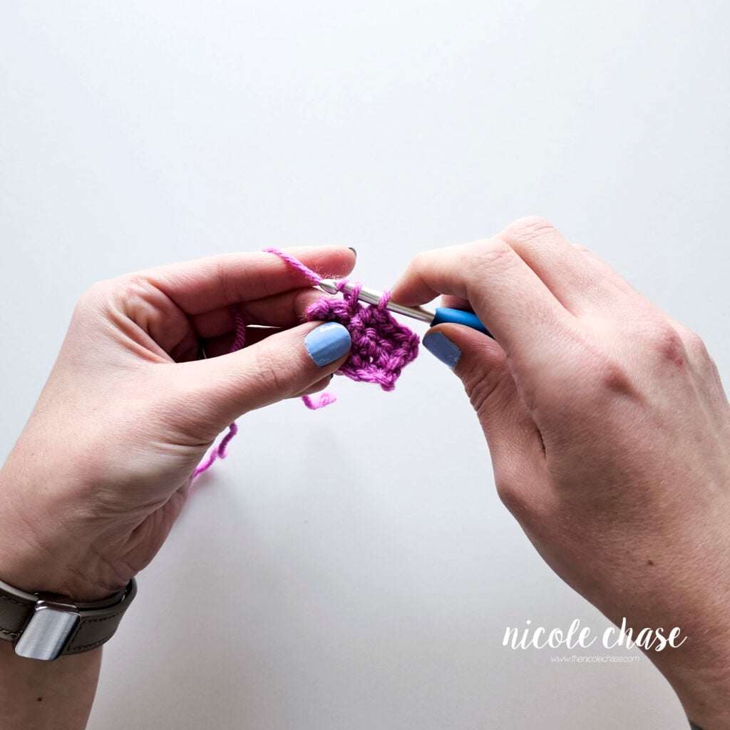 A close-up of hands holding a blue ergonomic crochet hook in the knife grip, initiating the next stitch. The purple yarn is pulled close, and the hook has the yarn over, ready to pull through the stitch. The tension hand stabilizes the previous small purple crochet swatch with the middle finger and thumb.