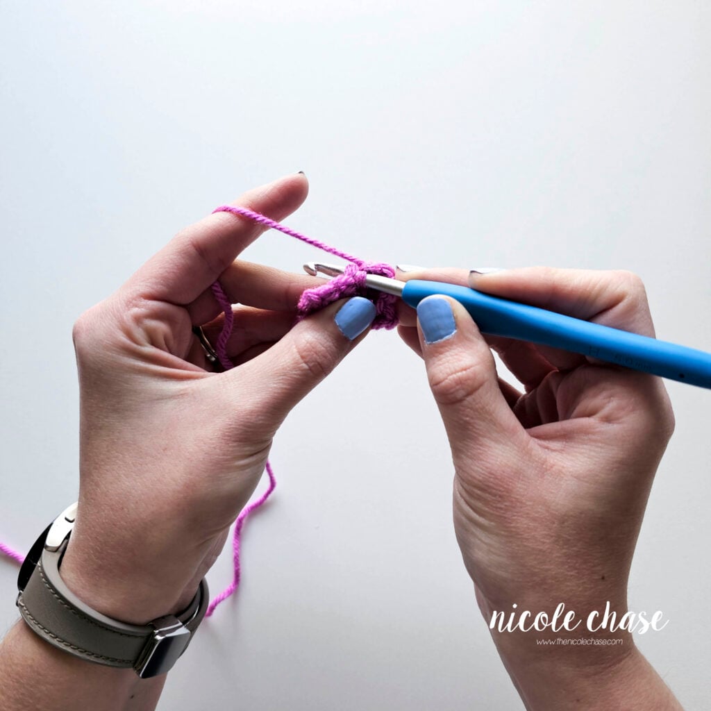 A close-up demonstrating the pencil grip for crocheting. The blue crochet hook is held between the thumb and index finger, while the tension hand holds a purple swatch and manages yarn flow. 