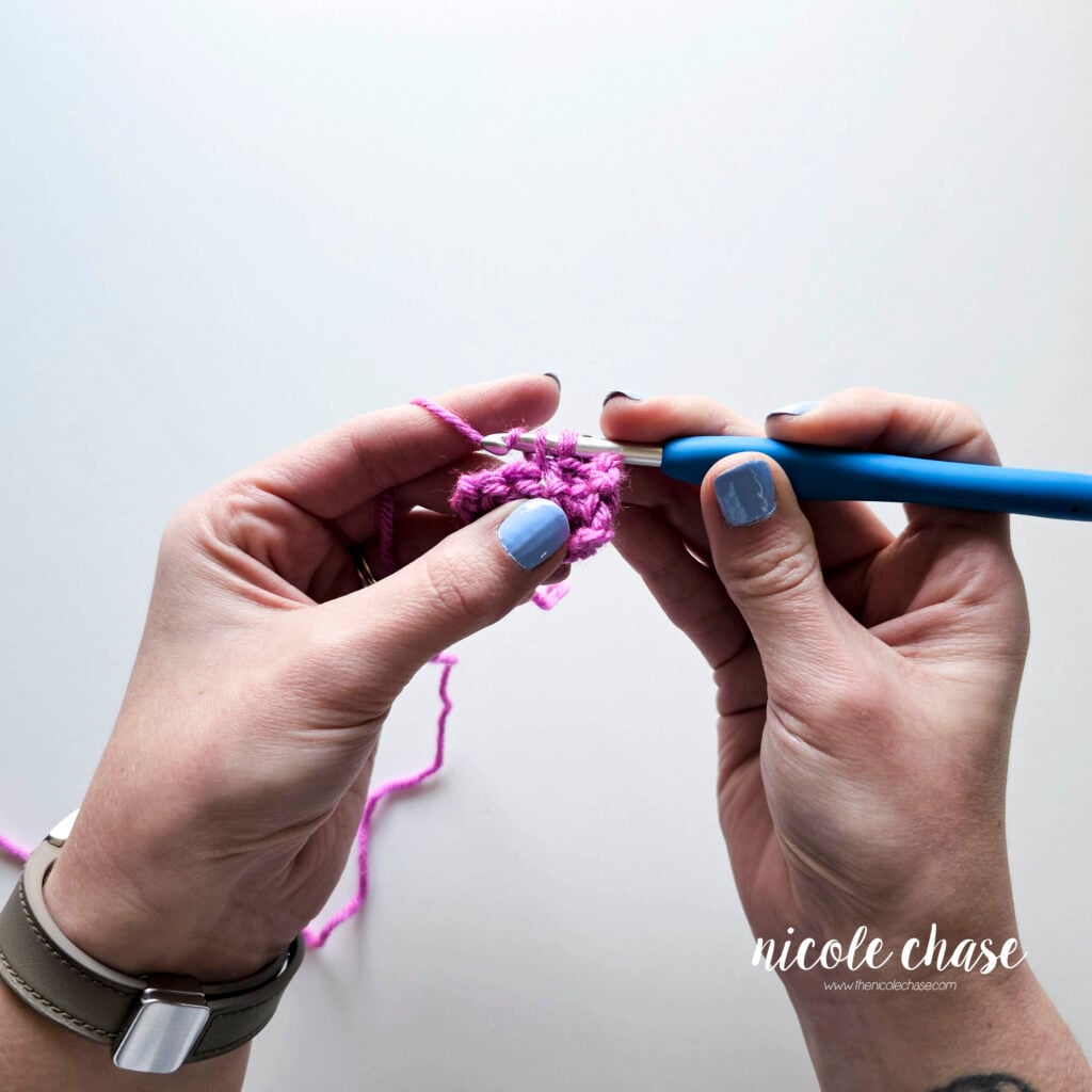 A tutorial image showing the pencil grip in action while working into a purple crochet stitch. The fingers of the right hand are positioned to pivot the blue hook while the left hand maintains yarn tension. 