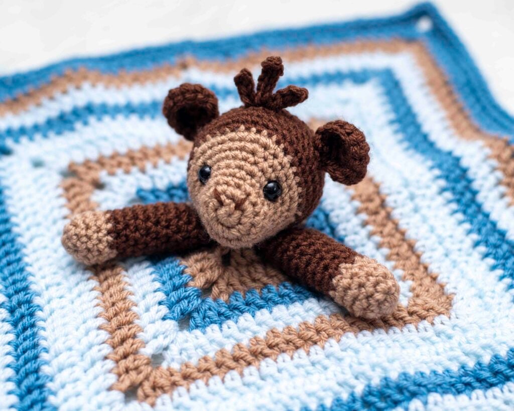 A close-up of a handmade amigurumi monkey love. The two toned brown and light beige monkey head with black safety eyes and a small tuft of hair is centered. The monkey's arms are spread across a square security blanket with alternating stripes of light blue, tan, and dark blue yarn.