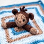 A close-up of a handmade amigurumi monkey love. The two toned brown and light beige monkey head with black safety eyes and a small tuft of hair is centered. The monkey's arms are spread across a square security blanket with alternating stripes of light blue, tan, and dark blue yarn.