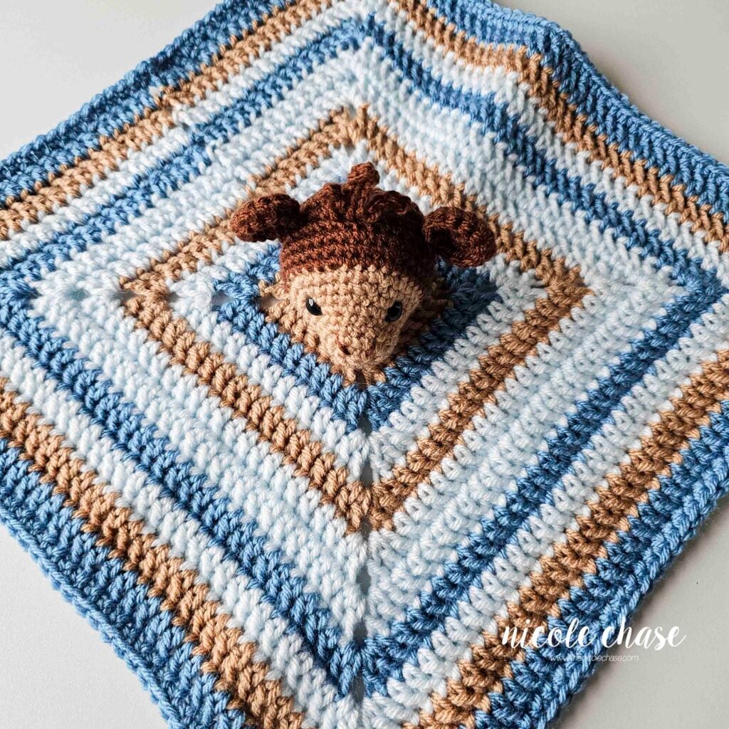 A top down view of the crochet monkey head having been sewn on to the center of the blue and brown crochet square lovey blanket. 