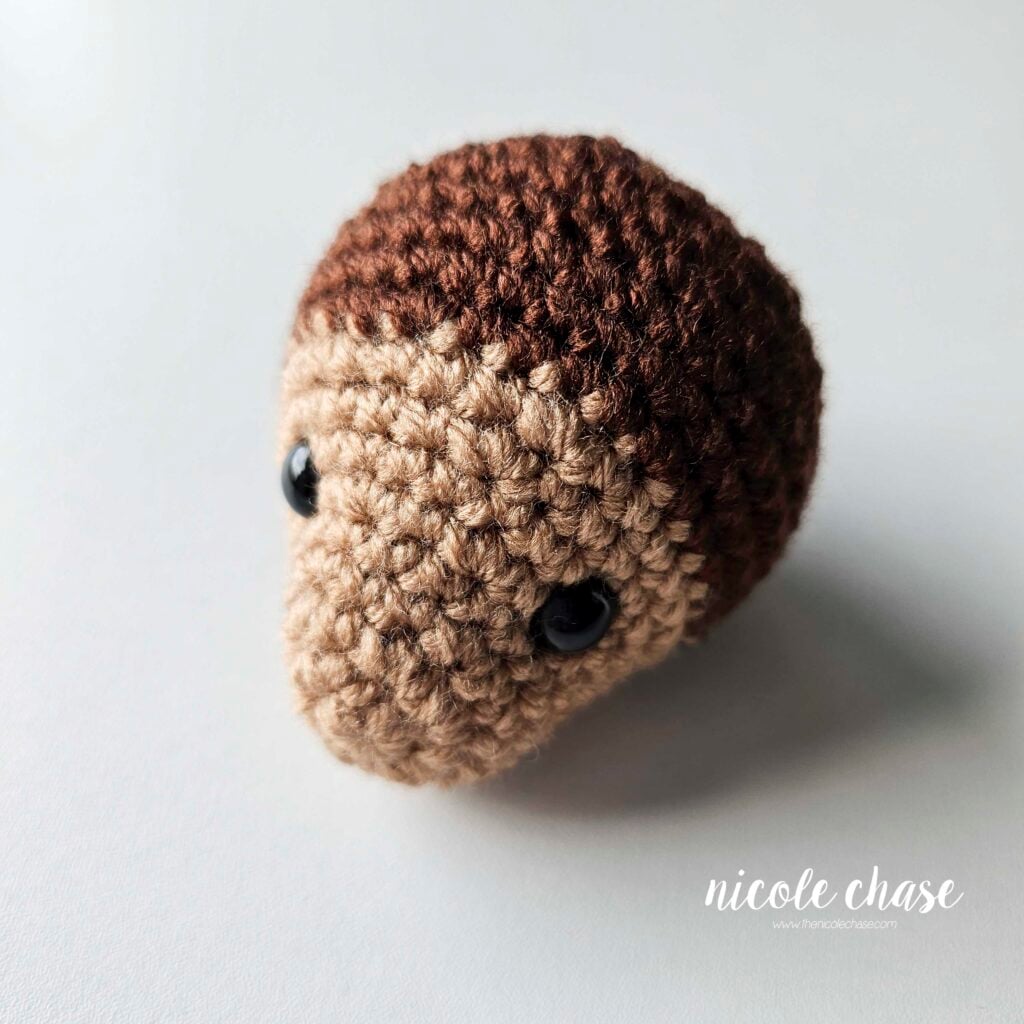 The finished head of the crochet monkey shown in a light brown yarn on the face and a dark brown yarn for the rest of the head, detailing the black safety eyes. The head is filled with stuffing and ready for assembling.