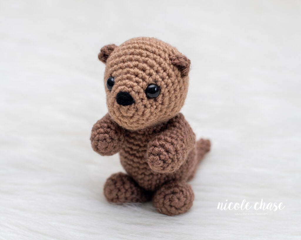 Side profile of a finished amigurumi otter from the free otter crochet pattern by Nicole Chase. This view highlights the 3D snout shaping, small rounded ears, and the attachment of the front and back limbs, sitting on a white textured background.