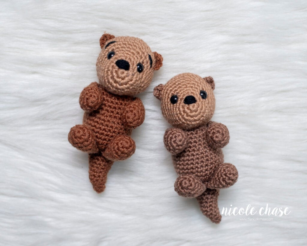 Two small amigurumi otters made from a free otter crochet pattern by Nicole Chase, shown in two different shades of brown yarn with black safety eyes, laying on a white faux fur background.