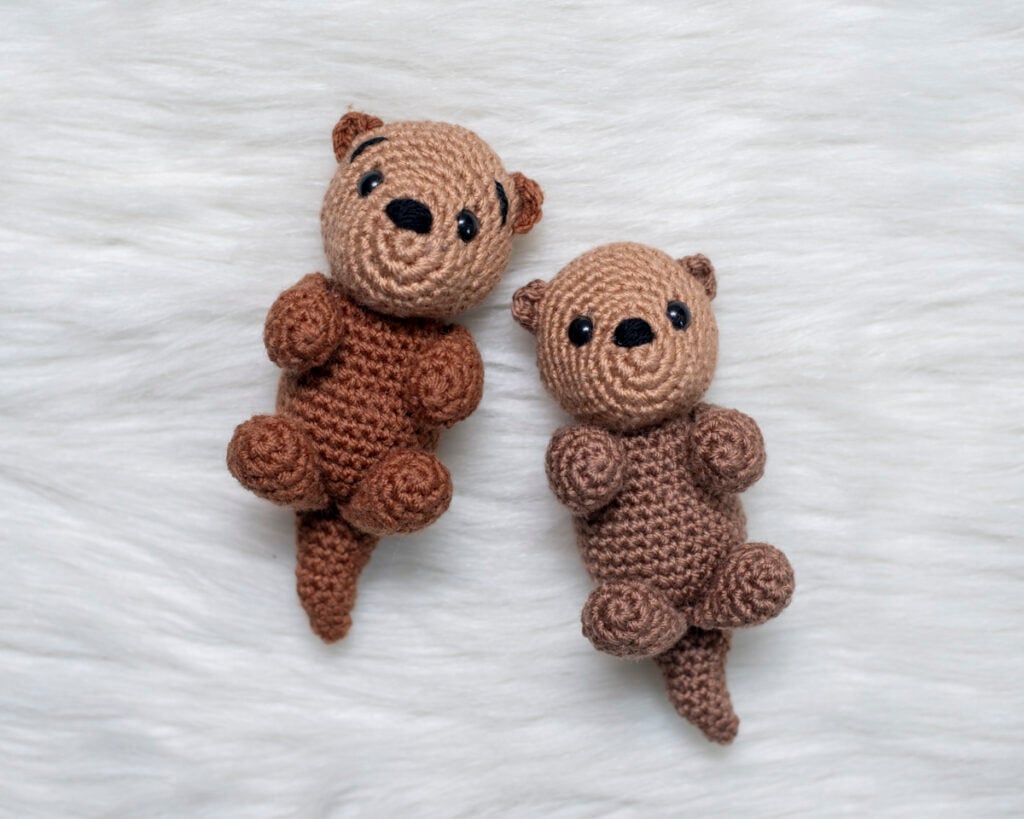 Two finished crochet sea otters, Ollie the Otter, showing the front and side view of the amigurumi project on a white fluffy surface.