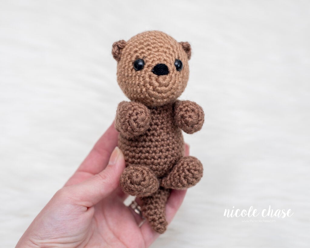 A hand holding a small, brown amigurumi otter to show its size. This free otter crochet pattern by Nicole Chase features a light brown head, black safety eyes, and a dark brown body, photographed against a white fur background.