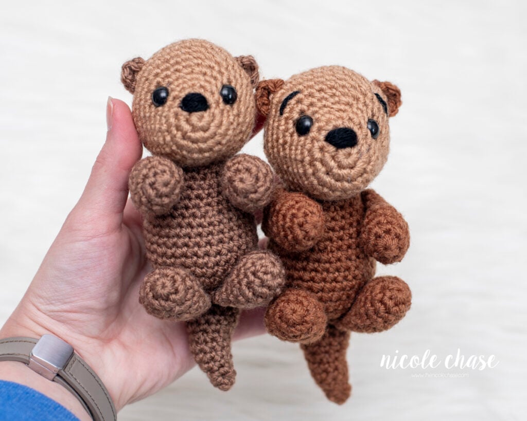 Two finished amigurumi otters held in one hand to show different brown yarn color combinations. This free otter crochet pattern demonstrates how Ollie looks in various shades, highlighting consistent shaping of the head, snout, and limbs.