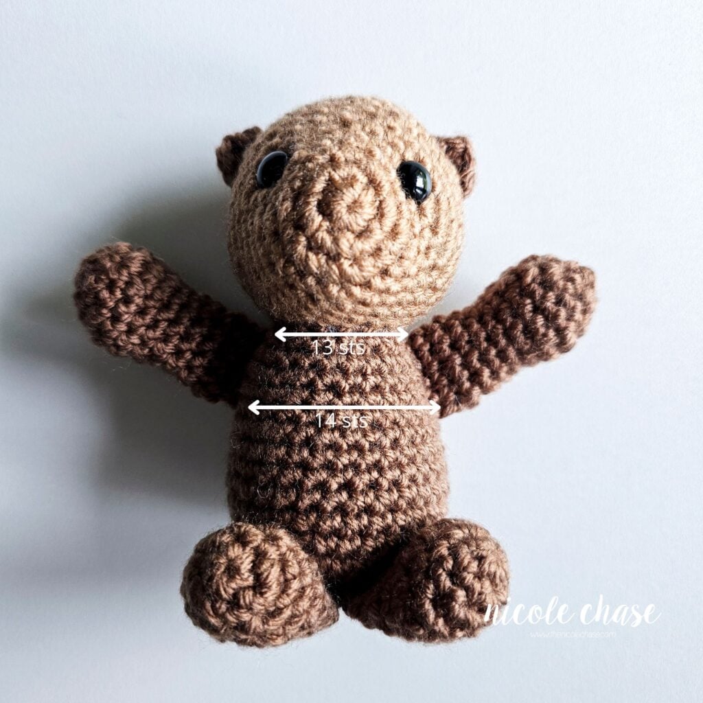 The crochet otter's arms are sewn into place and are sitting with it's arms open wide, with a graphic showing the number of stitches between each part of the arm.