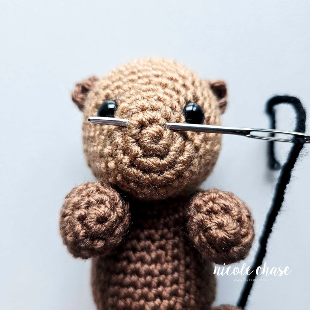 A yarn needle is threading black yarn through the front of the face of the crochet otter to embroider the nose.
