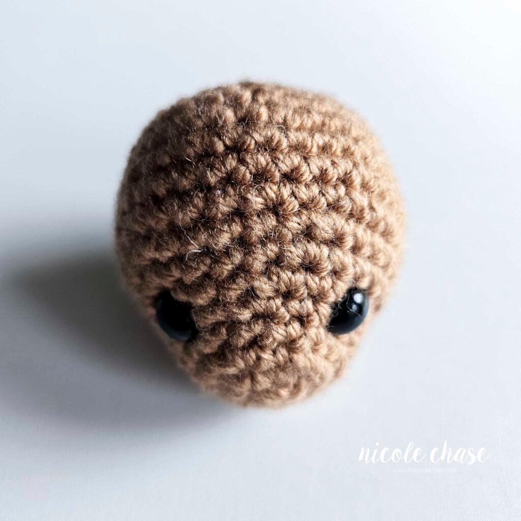 The finished crochet head of the amigurumi otter shown in a light brown yarn with black safety eyes. The head is filled with stuffing and waiting for assembly.